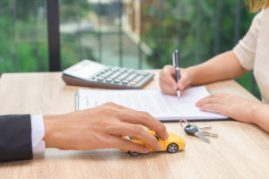 Woman signing car loan agreement contract with car key and calculator on wooden desk.